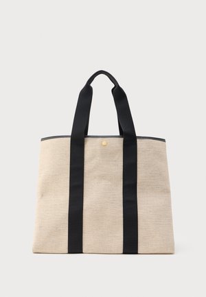 Beige stoffen draagtas met twee brede zwarte handvatten die van de bodem tot de bovenranden lopen, met een klein rond gouden embleem vlakbij de rits.