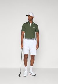 Polo verde con dettagli bianchi sul colletto, abbinato a pantaloni corti bianchi. Il modello tiene in mano una mazza da golf, indossando un cappellino bianco e scarpe sportive.