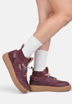 Ben iført hvide crew-sokker og bordeaux høje sneakers med farverige snørebånd og beige gummisåler på en ensfarvet baggrund.