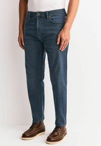 Blaue Jeans aus Denim mit schlanker Passform, Fünf-Taschen-Design und dezenten Nähten. Kombiniert mit braunen Schnürstiefeln für einen Kontrast.