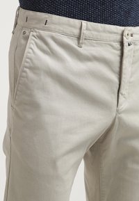 Pantaloni di cotone beige con tasche laterali, passanti per cintura e chiusura a bottone, indossati con un top in maglia blu scuro.