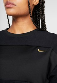 Musta huppari, jossa on teksturoitu yläpaneeli, pieni kultainen Nike-logo edessä, pyöreä kaula-aukko ja letitetty hiustyyli.