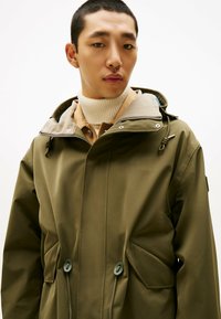 Wodoodporna kurtka w oliwkowym zielonym kolorze z kapturem ze ściągaczem, prążkowanym kołnierzem i dwoma zapinanymi na guziki kieszeniami. Posiada zamek błyskawiczny i minimalistyczny design.