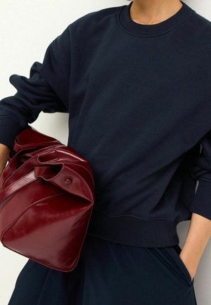Osoba ubrana w ciemnogranatowy sweter i spódnicę trzyma złożoną, błyszczącą głęboko czerwoną skórzaną torebkę na jednolitym jasnym tle.
