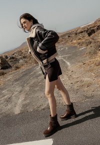 Stivali ankle in pelle marrone con un risvolto di lana, abbinati a una gonna nera e a un grande giubbotto in shearling grigio e nero, su un sentiero roccioso.