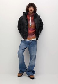 Młoda osoba z kręconymi włosami, ubrana w czarną kurtkę puchową, kolorowy sweter na zamek, luźne niebieskie dżinsy i beżowe sneakersy na jednolitym tle.