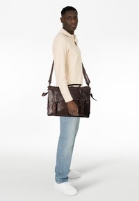 Bolso mensajero de cuero marrón con hebillas, un asa superior y una correa ajustable. Textura suave y forma rectangular.