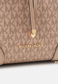 Gros plan sur un sac beige Michael Kors avec un motif MK répétitif, un logo doré et un cordon en cuir avec des accents dorés.