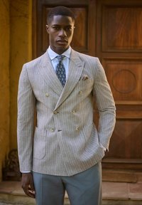 Blazer doppiopetto grigio chiaro con sottili righe bianche, due tasche e bottoni di legno, abbinato a una camicia azzurra e una cravatta fantasia.
