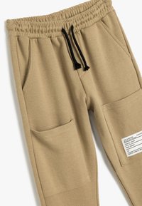 Sandfarvede sweatpants med tekstureret overflade, elastisk talje, sort snor og to forlommer. Inkluderer et mærke med tekst om stedet.