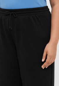 Fekete pamut melegítőnadrág húzózsinórral a derekán, sima textúrával és laza fazonnal. Kéz pihen a nadrág szárán.