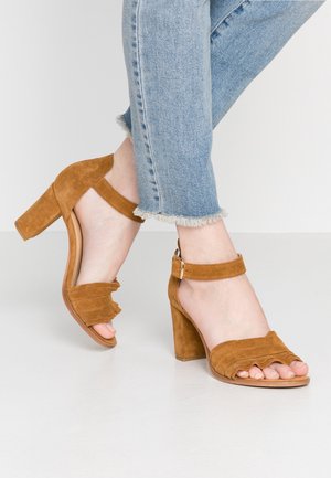 Brązowe zamszowe sandały na obcasie z blokowym obcasem, z falbaniastymi paskami i zapięciem wokół kostki. Matowa faktura i otwarty przód.