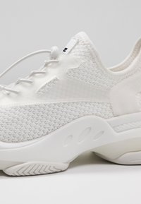 Chaussure de sport blanche avec tissu en maille texturé, lacets élastiques à arrêt et semelle épaisse avec rainures ovales et motif de la bande de roulement.