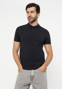 Homme portant un polo noir à manches courtes et un jean gris clair, debout les mains dans les poches, regardant sur le côté.