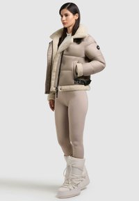 Beige Daunenjacke mit Schaffellkragen, schwarzen Akzenten und Knöpfen. Das Modell trägt beiges schmal geschnittenes Hosen und weiße isolierte Stiefel.