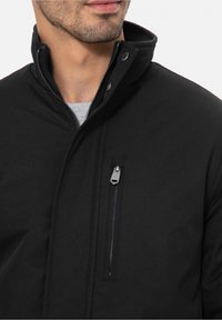 Sort jakke med høj krave, tekstureret stof, sidelomme med lynlås og trykknapper. Har en grå skjorte under.