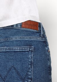 Blå denimjeans med en texturerad yta, som har en brun läderetikett från Wrangler på midjebandet och ett diamantformat sömnadsmönster på bakfickan.
