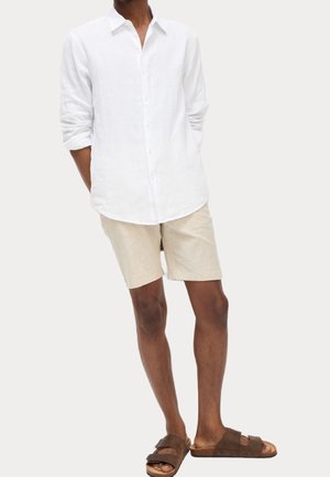 Mann trägt ein langärmeliges weißes Hemd mit Knöpfen, beige Shorts und braune Pantoletten, steht mit den Händen hinter dem Rücken.