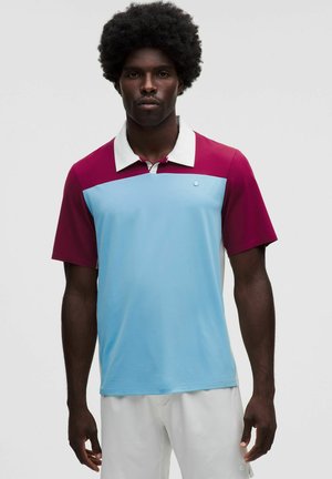Uomo con acconciatura afro che indossa una polo a maniche corte con spalle bordeaux e parte anteriore azzurro chiaro, in piedi davanti a uno sfondo uniforme.