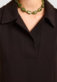 Czarny ubiór z kołnierzem i dekoracyjnym zielonym naszyjnikiem z koralików. Materiał wydaje się gładki, z delikatnymi detalami przeszyć.