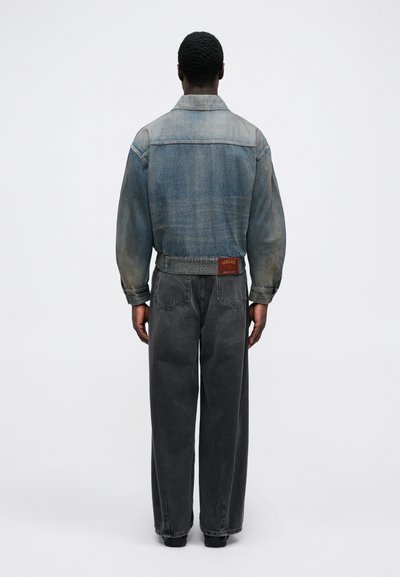 Denim jacket in light wash with a back yoke, paired with loose-fitting grey jeans. Notable branded leather patch at the waistband.