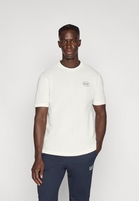 Λευκό T-shirt με κοντά μανίκια, στρογγυλή λαιμόκοψη και μικρό λογότυπο EA7 στο αριστερό μέρος του στήθους, σε συνδυασμό με μπλε σκούρο παντελόνι φόρμας. Λεία υφή.