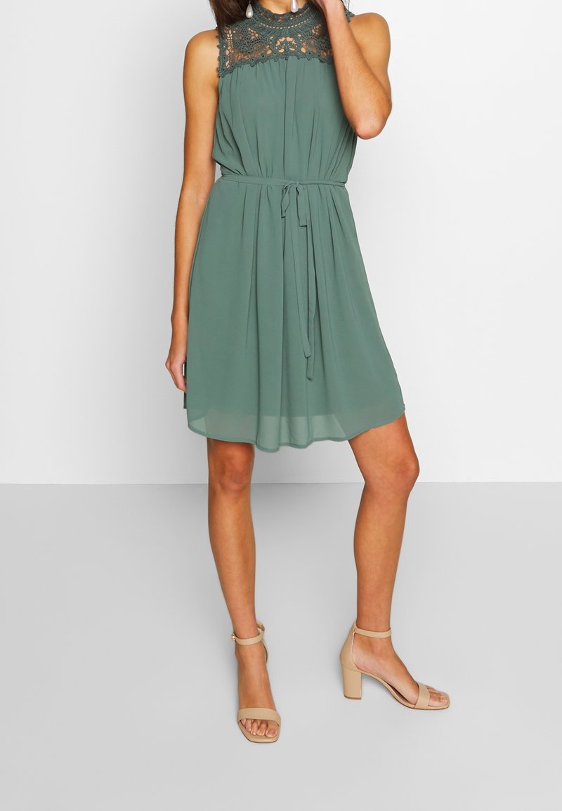 Robe verte sans manches avec empiècement en dentelle, taille nouée, ourlet au genou, portée avec des sandales beiges à talon bloc et bride autour de la cheville.