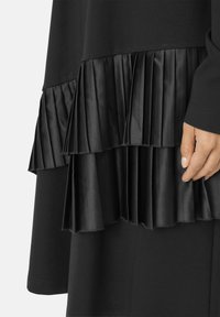 Sort kjole med et lagdelt stofdesign og satinfolder i kanten. Teksturerede og glatte sektioner står i visuel og taktil kontrast.