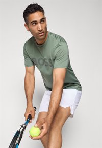 T-shirt de sport verte avec le logo "BOSS" en lettres foncées, associée à un short blanc. Tenant une balle de tennis et une raquette. Fond neutre.