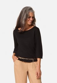 Zwarte, losse top met een wijde ronde hals en driekwartmouwen, gedragen met beige broek en een gouden kettingriem.
