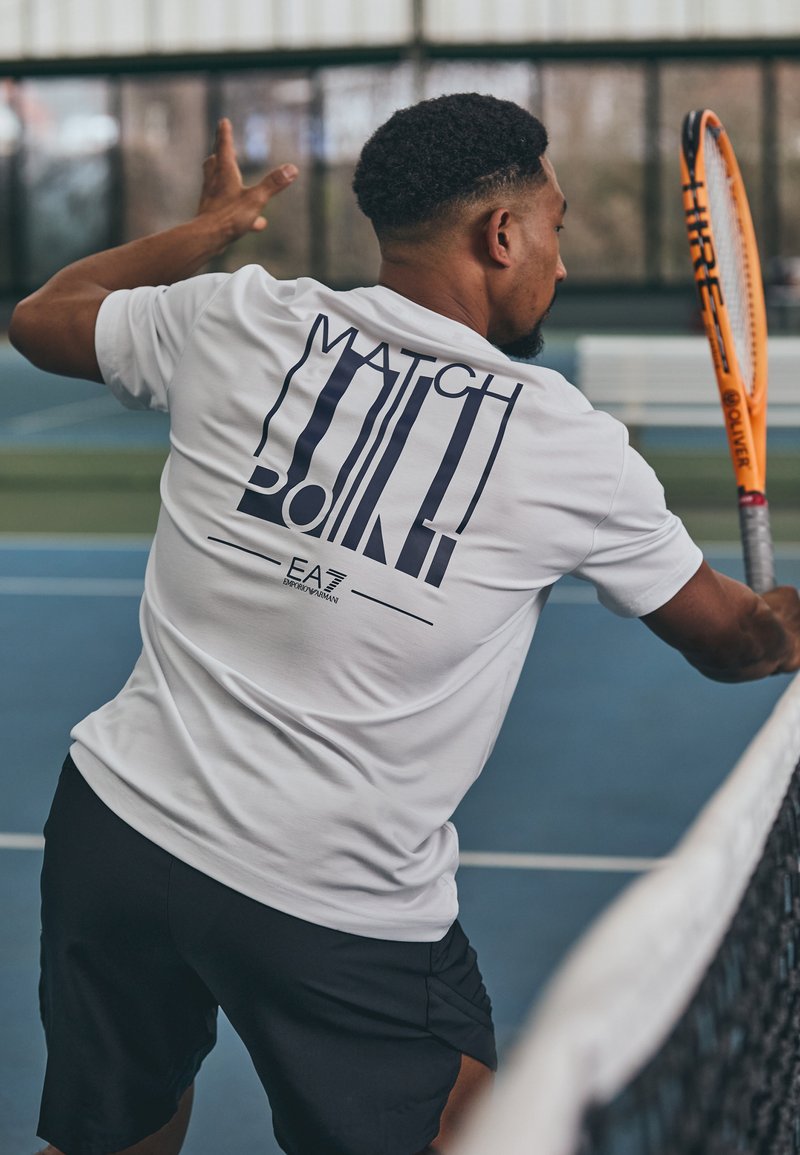 Balta trumpomis rankovėmis marškiniai su užrašu "MATCH POINT" tamsiai mėlyna spalva ant nugaros; derinama su juodomis šortais teniso aikštelėje.