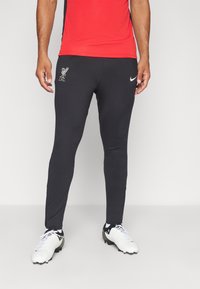 Sorte træningsbukser med slim fit, forsynet med et sølvfarvet Liverpool FC-logo, parret med hvide og sorte fodboldstøvler.