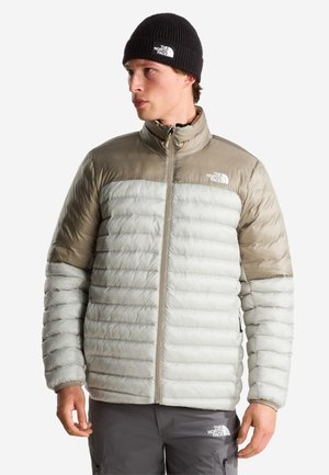 Junger Mann trägt eine grau-beige Daunenjacke, schwarze Mütze und graue Hose mit Markenlogos, schaut nach links vor einem schlichten Hintergrund.