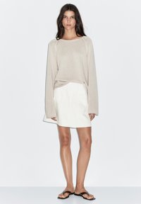 Beżowy sweter wykonany na drutach z długimi rękawami i szwami raglanowymi, w parze z białą spódnicą w kształcie litery A i czarnymi klapkami. Gładkie tło.