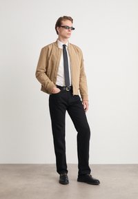 Veste bomber beige sur une chemise blanche et une cravate noire, avec un pantalon noir et des chaussures sombres. Les accessoires comprennent de petites lunettes de soleil noires.