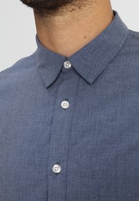 Camicia button-up in denim blu scuro con colletto tradizionale, due bottoni bianchi e una trama che appare liscia e leggermente strutturata.