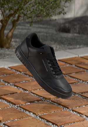Sneaker nera Tommy Hilfiger con lacci che fluttua sopra un pavimento in legno all'aperto, vicino a un albero.