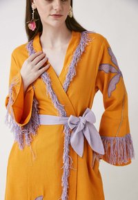 Bata naranja con ribete de flecos lavanda, ceñida a la cintura con un lazo. Presenta bordados florales en las mangas en acentos púrpura y dorado.