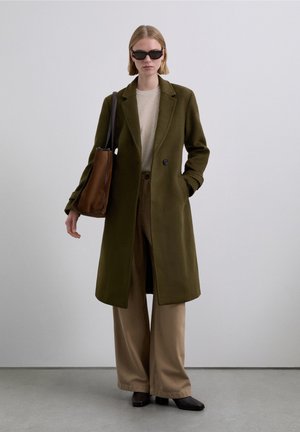 Abrigo de lana verde oliva con corte entallado, mangas largas, dos botones, combinado con un suéter beige y pantalones anchos beige, bolso de cuero marrón.
