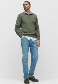 Mörkgrön tröja med dragkedjekrage, kombinerad med en ljusblå skjorta, ljusblå jeans och bruna skor. Minimalistisk design och avslappnad passform.