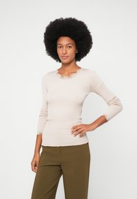 Moteris su afros šukuosena, dėvinti smėlio spalvos ilgarankovį marškinį su nėrinių krašte ir alyvuogių žalios spalvos kelnes, pozuoja, viena ranka pasidėjusi ant klubų.