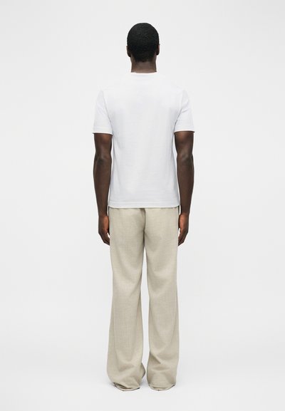 Άνδρας που στέκεται με την πλάτη προς τα εμπρός, φοράει απλό λευκό κοντομάνικο πουκάμισο και φαρδιά μπεζ παντελόνια μπροστά σε απλό λευκό φόντο.