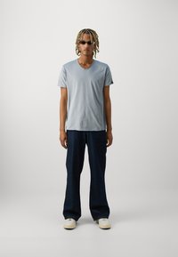 Tricou v-neck bleu deschis, pantaloni largi din denim închis, și adidași albi. Modelul are părul împletit și ochelari de soare.
