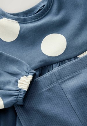 Blauwe katoenen sweatshirt met grote witte stippen, voorzien van geplooide mouwen; geribbelde blauwe broek met bijpassende textuur.