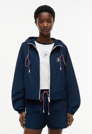 Mujer que lleva chaqueta con capucha azul marino y pantalones cortos con camiseta blanca, todos con cordones rojos y blancos y detalles del logo.