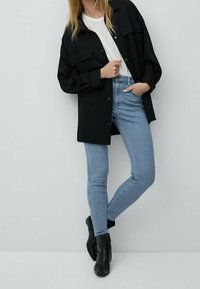 Mustat ylimitoitetut paidat, joissa on kaksi rintataskua, yhdistettyinä vaaleansinsisiin kapeisiin farkkuihin ja mustiin nilkkabootseihin, neutraalin taustan edessä.