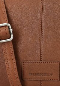 Borsa in pelle marrone con texture granulosa, dettagli cuciti e una fibbia in metallo argentato. Il nome del marchio "BURKELY" è impresso su una toppa.