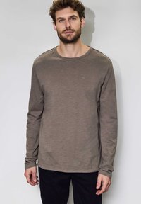 Långärmad skjorta i olivgrön bomull med rund hals, prydd med en liten logotyp på bröstet och textil med struktur. Bärs med mörka byxor.