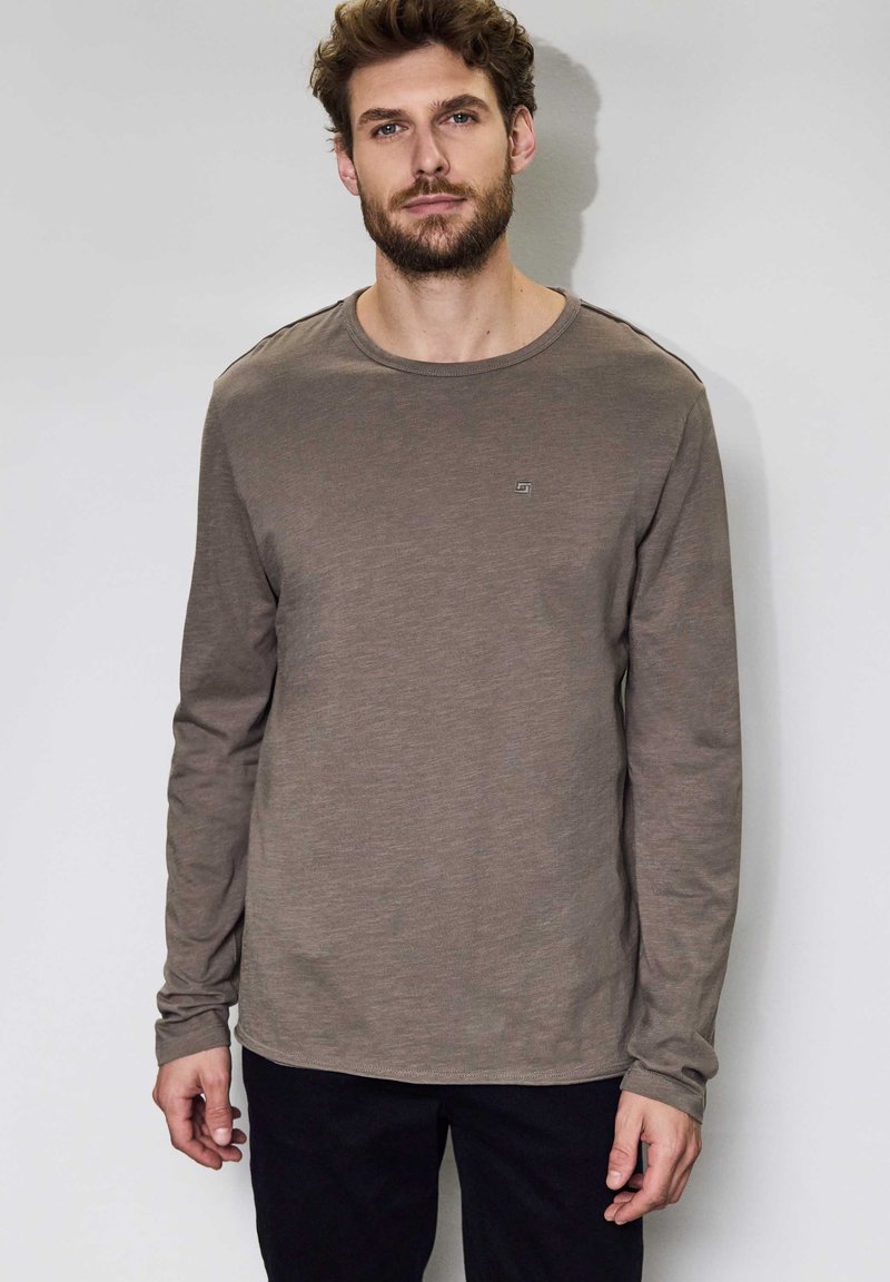 Långärmad skjorta i olivgrön bomull med rund hals, prydd med en liten logotyp på bröstet och textil med struktur. Bärs med mörka byxor.