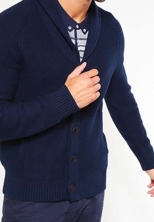 Mężczyzna noszący granatowy cardigan na guziki na paskowanej koszulce polo, poprawiający cardigan przy piersiach.